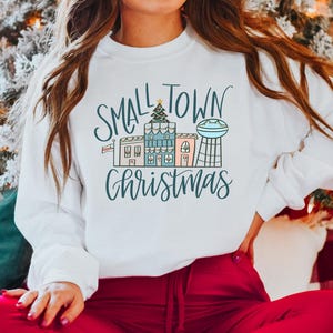 May include: White sweatshirt with a graphic design of a small town with houses and a water tower. The text "Small Town Christmas" is written in a cursive font.