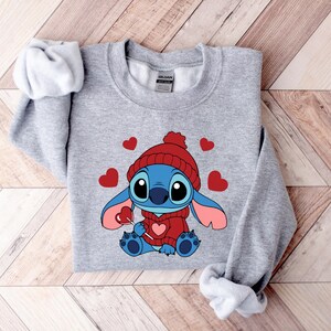 Pu&ograve; includere: Una felpa grigia con un personaggio dei cartoni animati con un cappello e un maglione rossi, che tiene in mano un lecca-lecca a forma di cuore. Il personaggio &egrave; blu con grandi occhi e orecchie rosa. Cuori rossi sparsi.