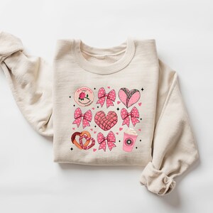 May include: A beige sweatshirt with a pink and brown graphic print of hearts, bows, and Mexican pastries. The text "La Gordita" is printed on the sweatshirt.