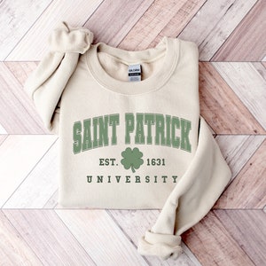 Puede incluir: Una sudadera beige con un tr&eacute;bol verde y el texto "Saint Patrick University Est. 1631" impreso en la parte delantera.