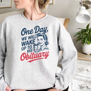 Puede incluir: Una sudadera gris claro con el texto "One Day, We Will Wake Up To His Obituary" en azul y rojo. El dise&ntilde;o incluye una ilustraci&oacute;n vintage de una mujer sosteniendo una taza de caf&eacute;.