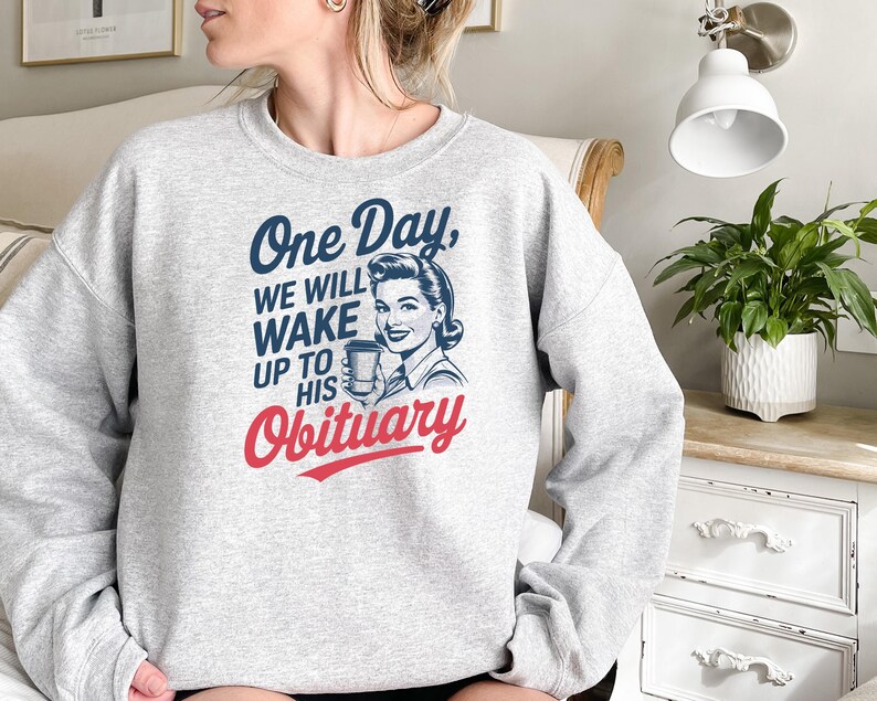 Puede incluir: Una sudadera gris claro con el texto "One Day, We Will Wake Up To His Obituary" en azul y rojo. El dise&ntilde;o incluye una ilustraci&oacute;n vintage de una mujer sosteniendo una taza de caf&eacute;.