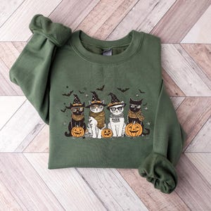 May include: Green sweatshirt with a graphic of four cats wearing witch hats and holding pumpkins. The cats are wearing glasses and have different colored fur.