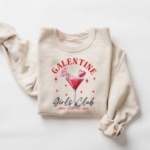 May include: A beige sweatshirt with the text "Galentine's Girls Club" in red and black lettering. The design also includes a pink cocktail with a heart-shaped garnish and stars.