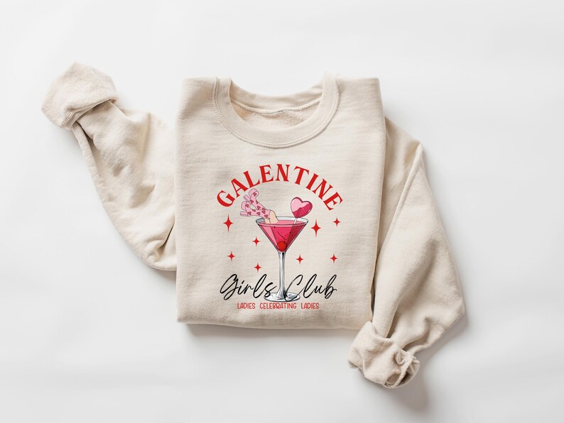 May include: A beige sweatshirt with the text "Galentine's Girls Club" in red and black lettering. The design also includes a pink cocktail with a heart-shaped garnish and stars.