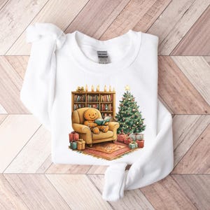 May include: White sweatshirt with a gingerbread person sitting in a chair reading a book next to a Christmas tree and a bookshelf with candles. The gingerbread person is wearing a red and white striped shirt and a brown hat.