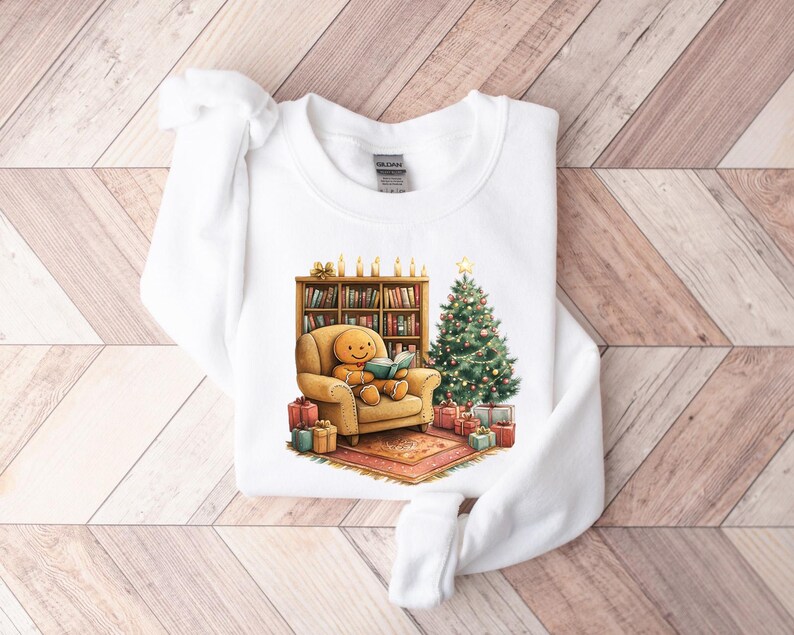 May include: White sweatshirt with a gingerbread person sitting in a chair reading a book next to a Christmas tree and a bookshelf with candles. The gingerbread person is wearing a red and white striped shirt and a brown hat.