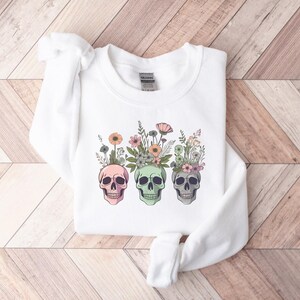 May include: White sweatshirt with a graphic of three colorful skulls with floral arrangements on top. The skulls are pink, green, and white.