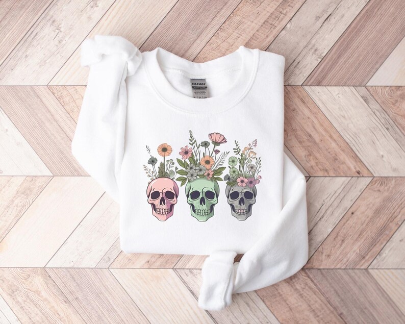 May include: White sweatshirt with a graphic of three colorful skulls with floral arrangements on top. The skulls are pink, green, and white.