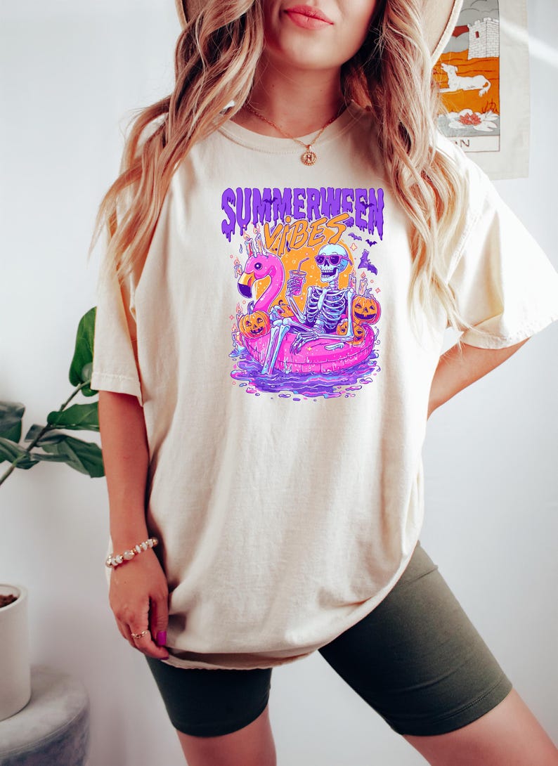 May include: Cream-colored t-shirt with a colorful graphic. The graphic features a skeleton on a pink flamingo floatie, with the text "SUMMERWEEN VIBES" in purple and orange. The shirt is a casual, relaxed fit.