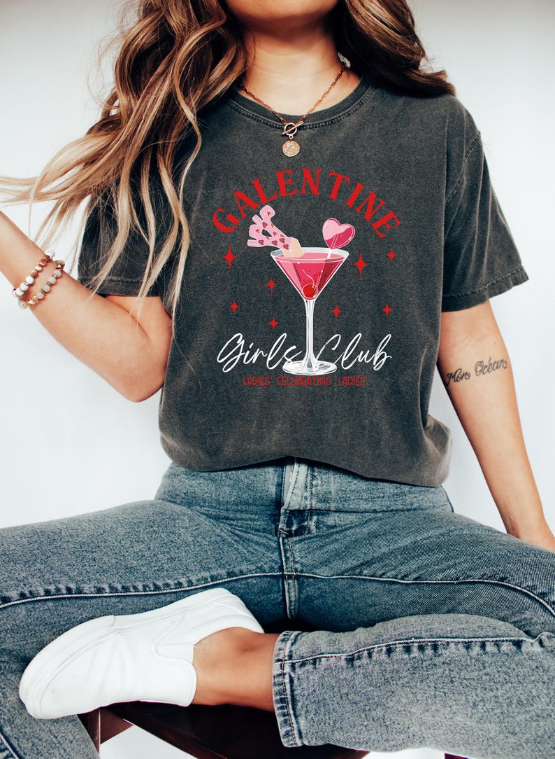 May include: A dark gray t-shirt with a red graphic design that says "Galentine's Girls Club" with a pink cocktail glass and hearts.