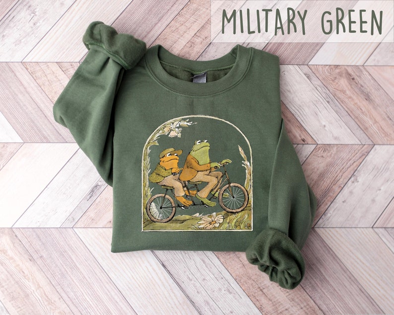 May include: A green sweatshirt with a graphic of two frogs riding a bicycle. The frogs are wearing clothes and the bicycle has a basket. The text "MILITARY GREEN" is printed in white at the top of the image.