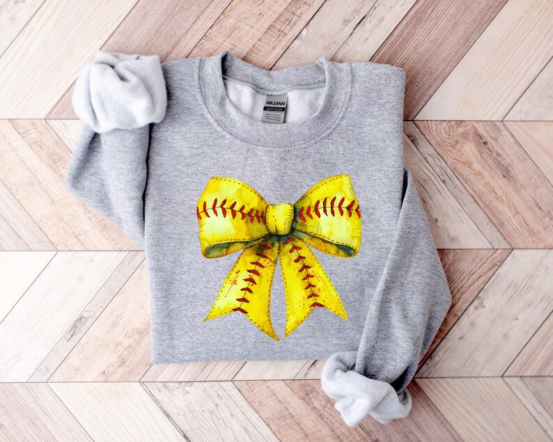 May include: A gray sweatshirt with a yellow and red softball bow design. The bow is tied with a red ribbon and has white stitching.