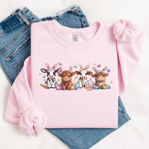 May include: Pink sweatshirt with a graphic of four cows wearing bunny ears and holding Easter eggs. The cows are black and white, brown and white, and brown.
