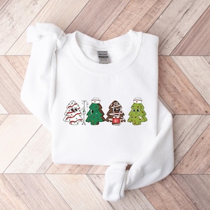 May include: White sweatshirt with a graphic of four Christmas trees dressed as nurses. Each tree has a red cross, a nurse's cap, and a different colored face.