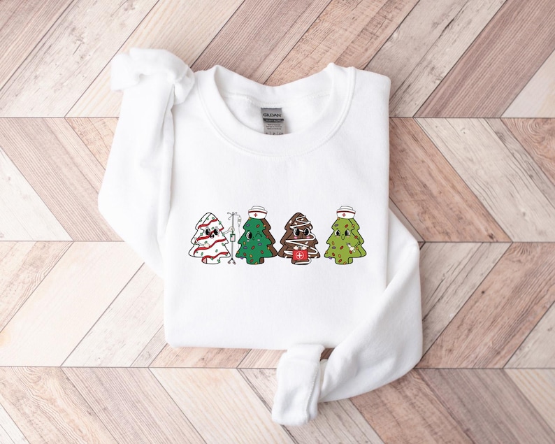 May include: White sweatshirt with a graphic of four Christmas trees dressed as nurses. Each tree has a red cross, a nurse's cap, and a different colored face.