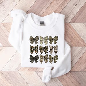 May include: White sweatshirt with a pattern of nine camouflage bows in shades of green, brown, and beige.