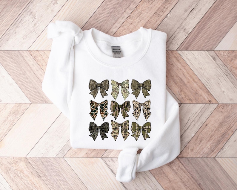 May include: White sweatshirt with a pattern of nine camouflage bows in shades of green, brown, and beige.