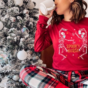 Puede incluir: Una mujer con pijama rojo con un gr&aacute;fico de esqueleto blanco y el texto "Spooky Nurse". Est&aacute; sosteniendo una taza blanca y sentada frente a un &aacute;rbol de Navidad cubierto de nieve.