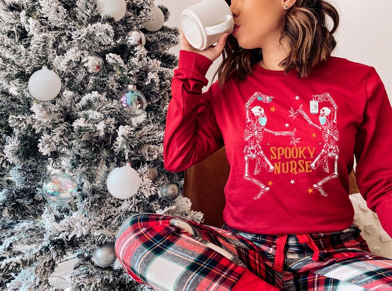 Puede incluir: Una mujer con pijama rojo con un gr&aacute;fico de esqueleto blanco y el texto "Spooky Nurse". Est&aacute; sosteniendo una taza blanca y sentada frente a un &aacute;rbol de Navidad cubierto de nieve.