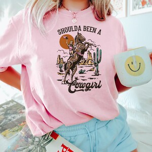 以下が含まれることがあります： 砂漠の風景の中で馬に乗っている女性のグラフィックがプリントされたピンクのTシャツ。Tシャツには「Shoulda Been a Cowgirl」という文字がプリントされています。