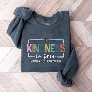 May include: A dark gray sweatshirt with the text "KINDNESS is free" in colorful letters inside a white rectangle. The text "SPRINKLE IT EVERYWHERE" is below the rectangle.