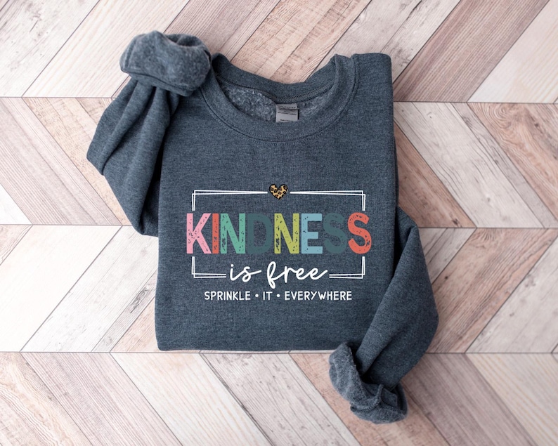May include: A dark gray sweatshirt with the text "KINDNESS is free" in colorful letters inside a white rectangle. The text "SPRINKLE IT EVERYWHERE" is below the rectangle.