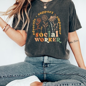 May include: A dark gray t-shirt with a graphic design featuring the words "social worker" in colorful letters surrounded by flowers and the words "advocate", "support", and "empower".