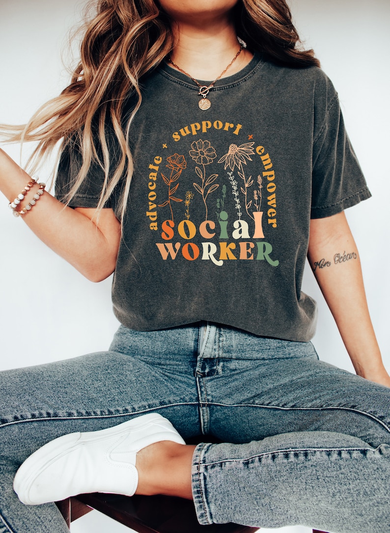 May include: A dark gray t-shirt with a graphic design featuring the words "social worker" in colorful letters surrounded by flowers and the words "advocate", "support", and "empower".