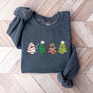 May include: A dark gray sweatshirt with a graphic of four Christmas trees dressed as nurses. The trees are wearing white nurse hats and have red crosses on their trunks.