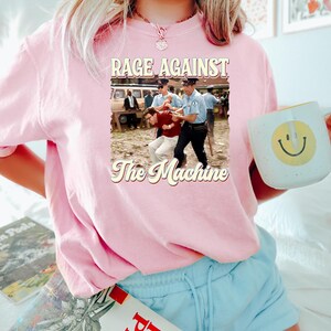 K&ouml;nnte beinhalten: Ein pinkfarbenes T-Shirt mit einer Grafik im Vintage-Stil einer Band namens "Rage Against The Machine". Die Grafik zeigt zwei M&auml;nner auf einem Schwarzwei&szlig;foto, wobei einer von dem anderen verhaftet wird.