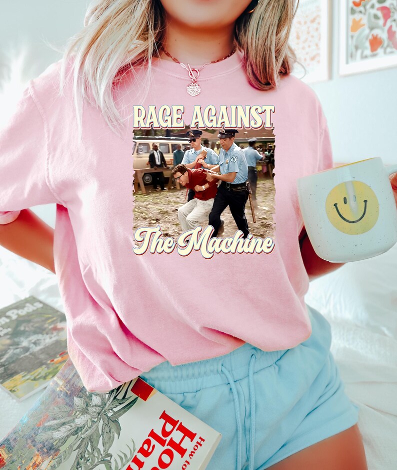 K&ouml;nnte beinhalten: Ein pinkfarbenes T-Shirt mit einer Grafik im Vintage-Stil einer Band namens "Rage Against The Machine". Die Grafik zeigt zwei M&auml;nner auf einem Schwarzwei&szlig;foto, wobei einer von dem anderen verhaftet wird.