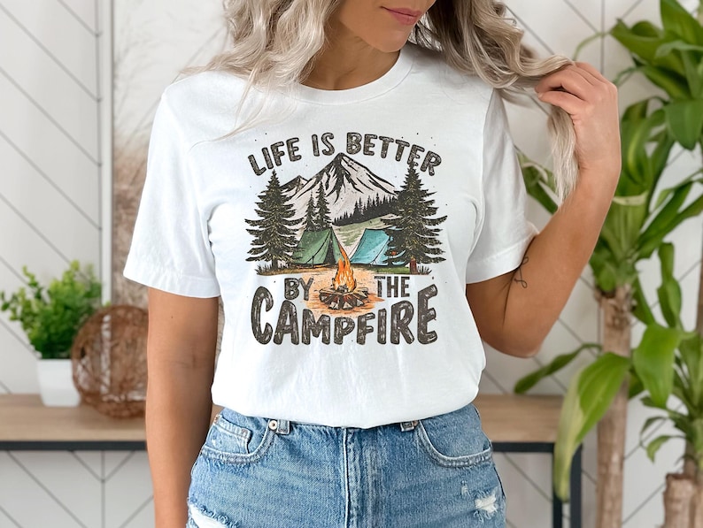 K&ouml;nnte beinhalten: Wei&szlig;es T-Shirt mit einem Grafikdesign einer Feuerstellenszene mit Bergen, B&auml;umen und Zelten. Der Text "Life is better by the campfire" ist auf das Shirt gedruckt.