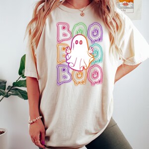 Puede incluir: Una camiseta blanca con un gr&aacute;fico de arco&iacute;ris de ne&oacute;n de la palabra "BOO" con un fantasma en el medio. El fantasma es blanco, tiene dos ojos y est&aacute; haciendo un signo de paz con ambas manos.