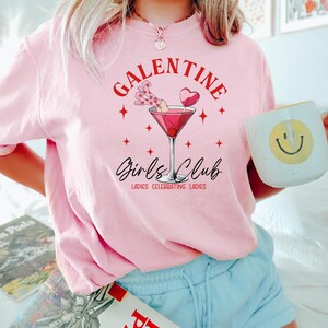 May include: Pink t-shirt with the text "Galentine's Girls Club" and a martini glass with a cherry and a heart-shaped lollipop. The text below the martini glass reads "Ladies Celebrating Ladies."