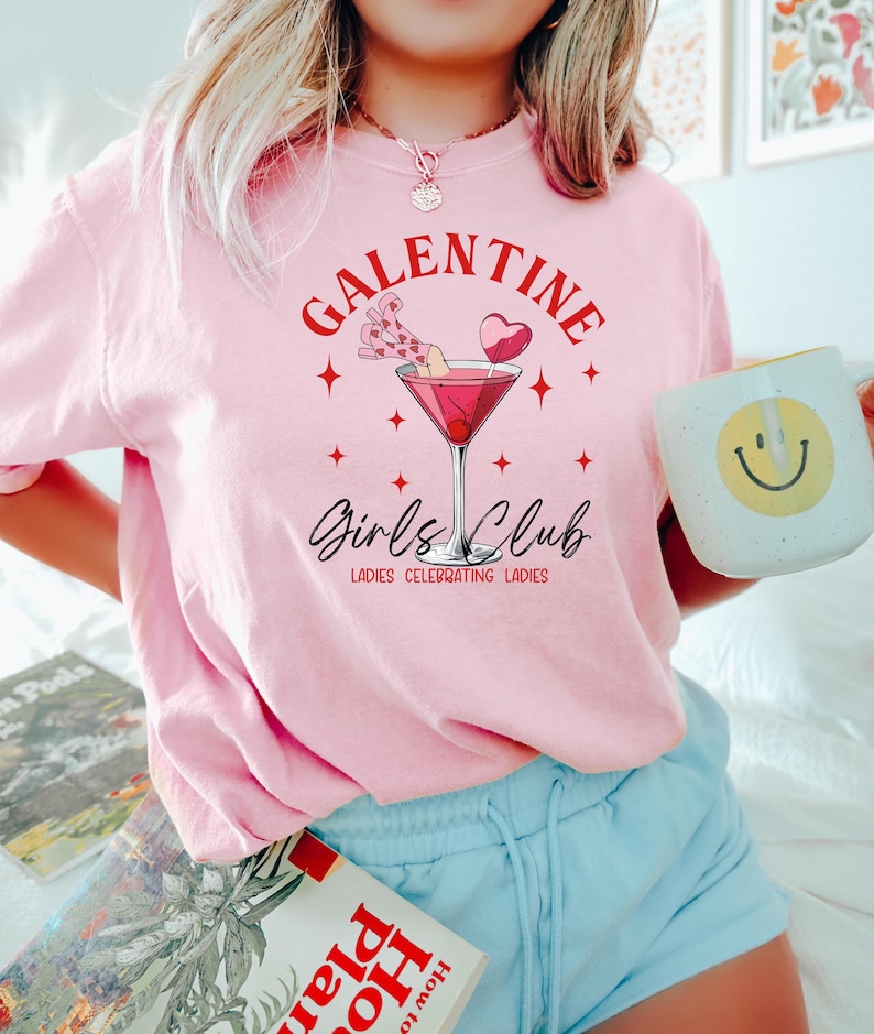 May include: Pink t-shirt with the text "Galentine's Girls Club" and a martini glass with a cherry and a heart-shaped lollipop. The text below the martini glass reads "Ladies Celebrating Ladies."