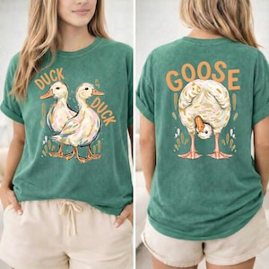 Puede incluir: Camiseta verde con un gr&aacute;fico de pato en la parte delantera y un gr&aacute;fico de ganso en la parte trasera. La parte delantera de la camiseta presenta dos patos con las palabras "DUCK DUCK" en naranja. La parte trasera de la camiseta presenta un ganso con la palabra "GOOSE" en naranja.