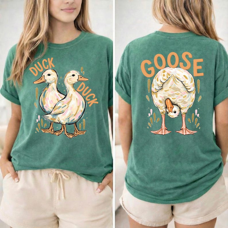 Puede incluir: Camiseta verde con un gr&aacute;fico de pato en la parte delantera y un gr&aacute;fico de ganso en la parte trasera. La parte delantera de la camiseta presenta dos patos con las palabras "DUCK DUCK" en naranja. La parte trasera de la camiseta presenta un ganso con la palabra "GOOSE" en naranja.