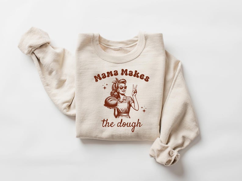 May include: A beige sweatshirt with a brown graphic of a woman holding a loaf of bread and the text "Mama Makes the Dough".