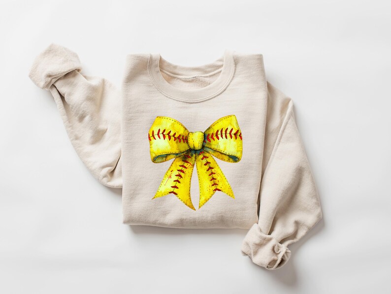 May include: A beige sweatshirt with a yellow and red softball bow design.