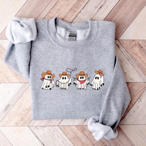 May include: A heather gray sweatshirt featuring a graphic of four cartoon ghosts dressed as cowboys. Each ghost wears a cowboy hat, with bandanas and other cowboy accessories. The design is centered on the front of the sweatshirt.