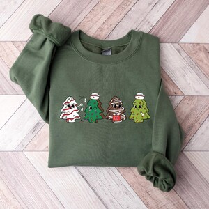 May include: A green sweatshirt with a Christmas tree design featuring four nurses wearing Santa hats. The trees are decorated with candy canes, ornaments, and medical symbols.
