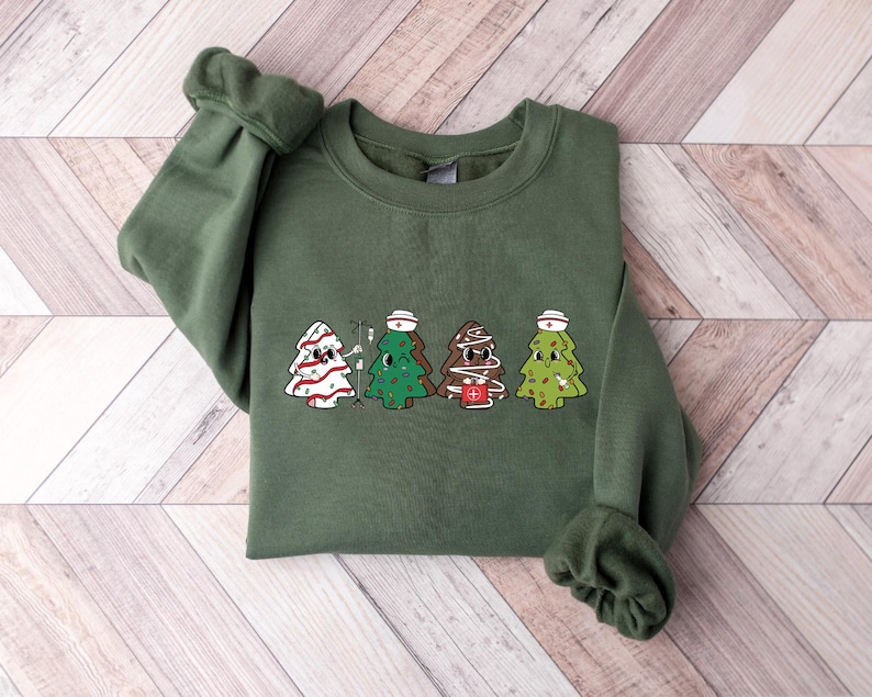 May include: A green sweatshirt with a Christmas tree design featuring four nurses wearing Santa hats. The trees are decorated with candy canes, ornaments, and medical symbols.