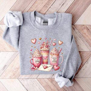 May include: Gray sweatshirt with a Valentine's Day design featuring three mugs of hot chocolate with whipped cream and hearts. The mugs have the text "XOXO", "Hugs and Kisses", and a heart.