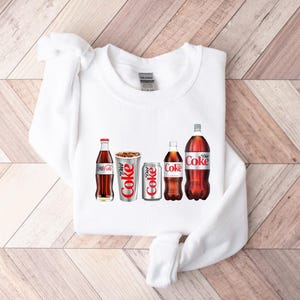 May include: White sweatshirt featuring a graphic of five Coca-Cola bottles and cans. The graphic includes a regular Coca-Cola bottle, a Diet Coke can, a Coca-Cola can, a Diet Coke bottle, and a Coca-Cola bottle.