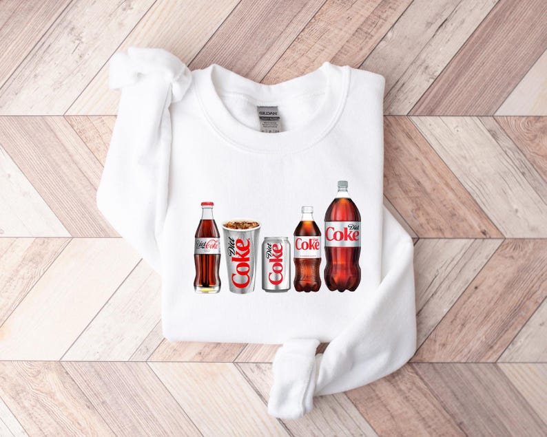 May include: White sweatshirt featuring a graphic of five Coca-Cola bottles and cans. The graphic includes a regular Coca-Cola bottle, a Diet Coke can, a Coca-Cola can, a Diet Coke bottle, and a Coca-Cola bottle.