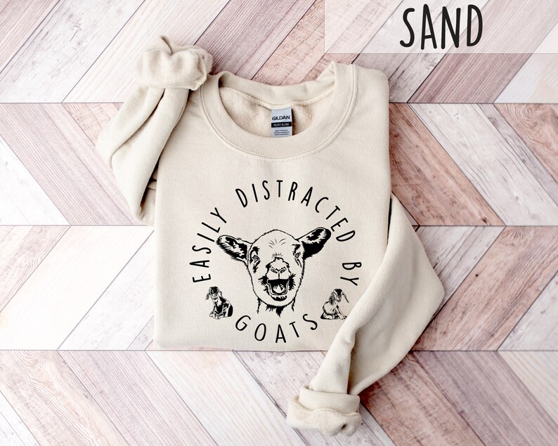 May include: A beige sweatshirt with the text "Easily Distracted By Goats" in black lettering and a goat illustration.