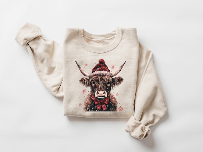 May include: A beige sweatshirt with a brown and white highland cow wearing a red Santa hat and a red bow tie. The cow is surrounded by small pink snowflakes.