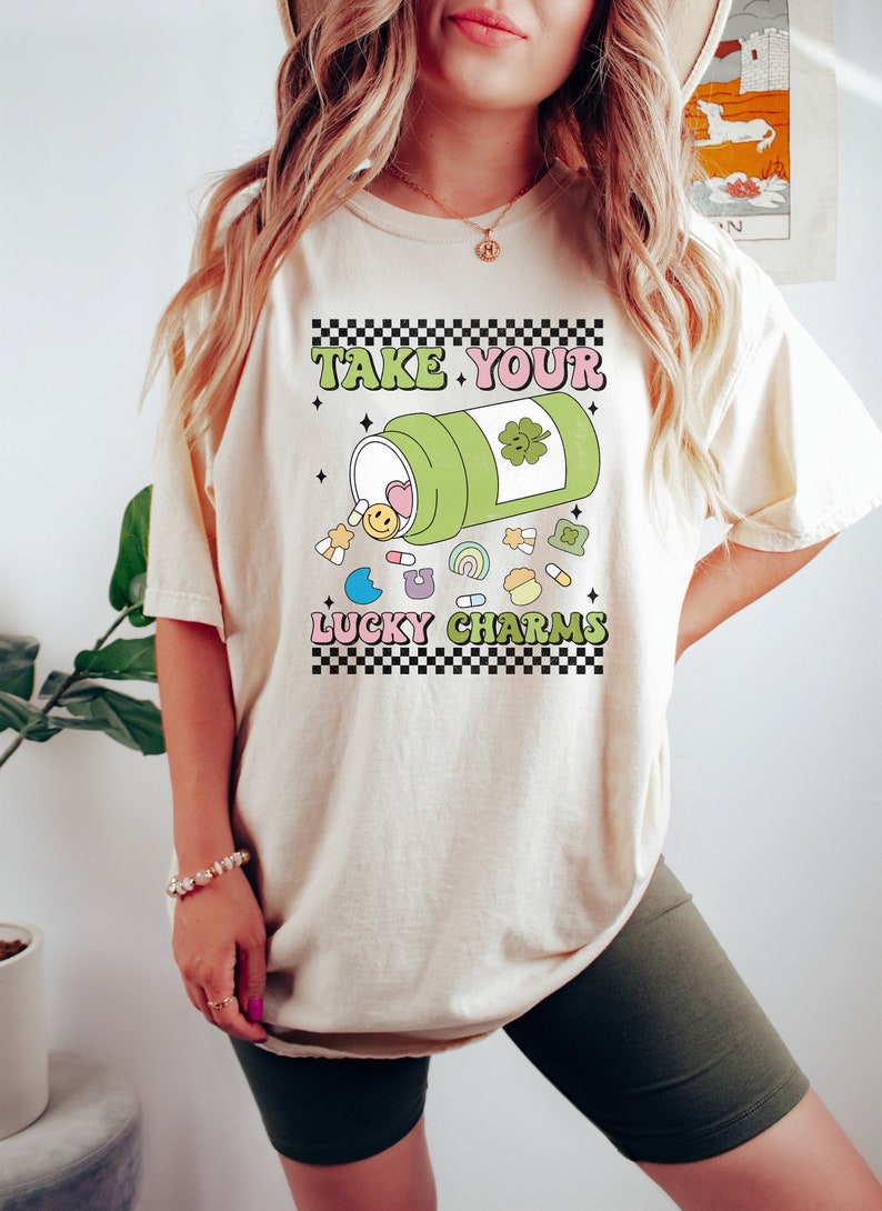 May include: A white t-shirt with a graphic of a green pill bottle with a four-leaf clover on it. The pill bottle is spilling out colorful cartoon charms, including a heart, a smiley face, a star, a horseshoe, a rainbow, and a clover. The text "TAKE YOUR LUCKY CHARMS" is printed in black and white checkered font.