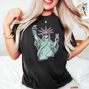 Może przedstawiać: Czarny t-shirt z grafiką przedstawiającą Statuę Wolności w okularach przeciwsłonecznych i z bańką z gumy do żucia. Statua jest w kolorze jasnoniebieskim z r&oacute;żowym obrysem. Koszulka jest w ciemnym kolorze.